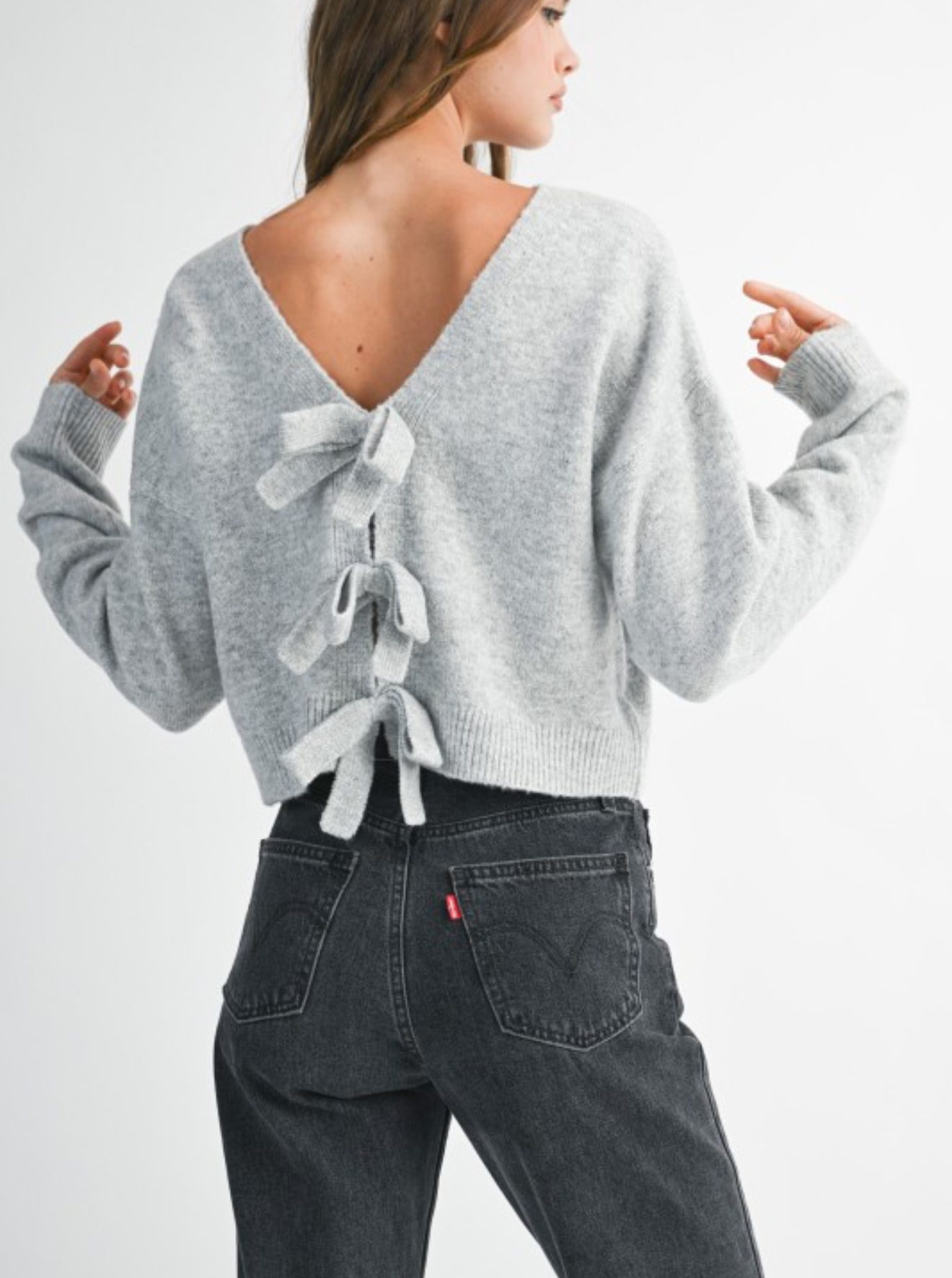 Woman wearing a gray sweater with ribbon bow detail and dark jeans on a white background